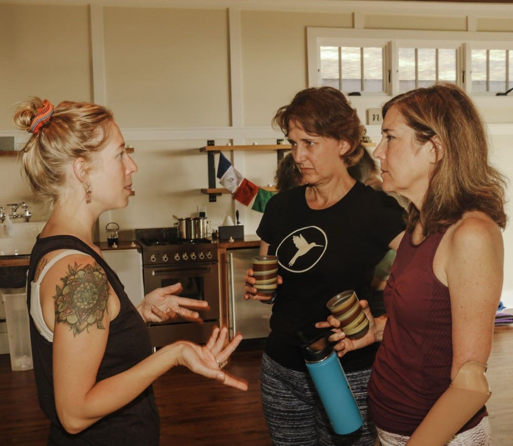 Three women are talking in a kitchen.